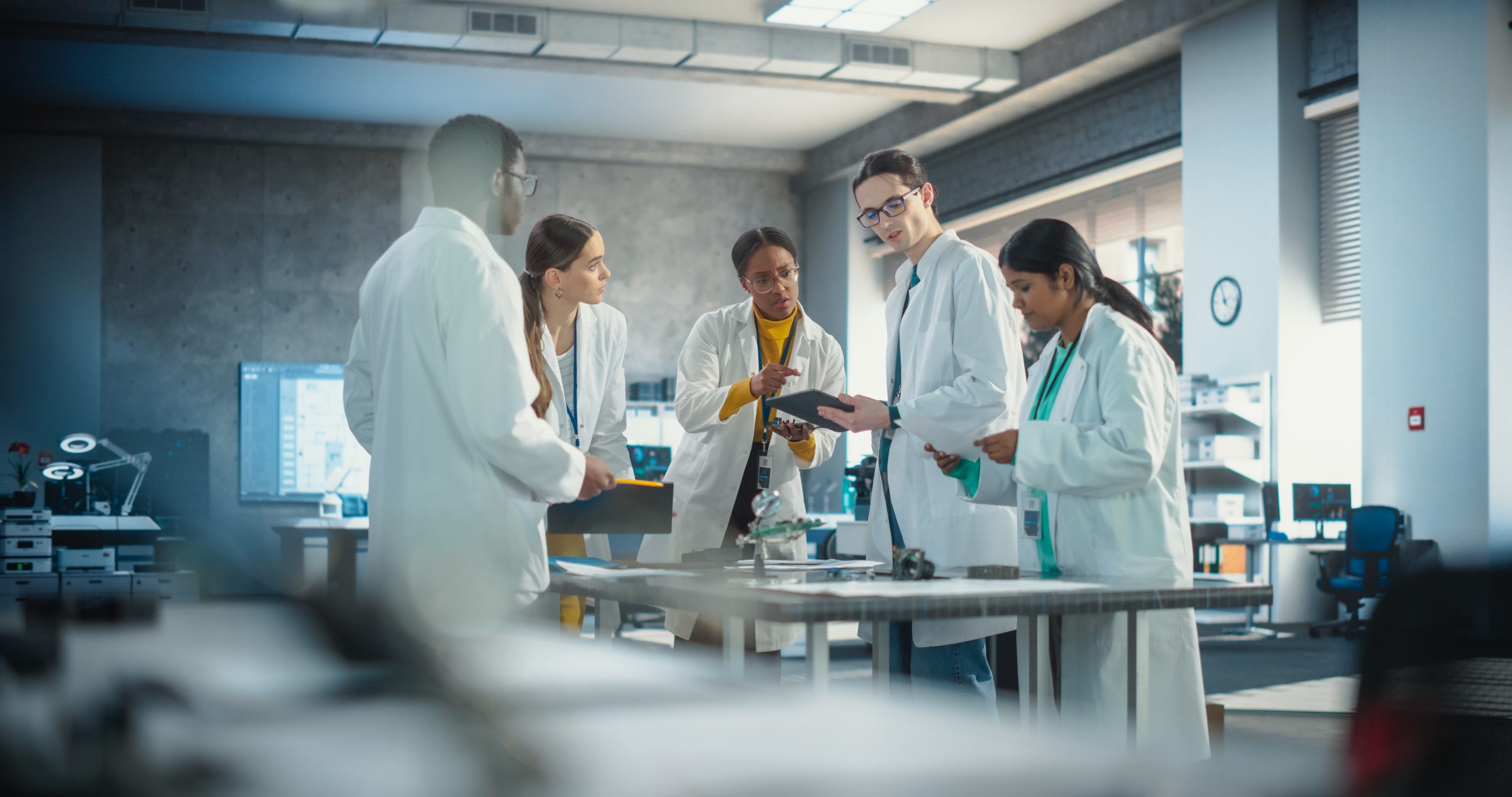 A group of people wearing personal protective equipment, such as lab coats and safety glasses, stand around a work table in an engineering or robotics-type lab, sharing a diversity of ideas about prototypes and documents.
