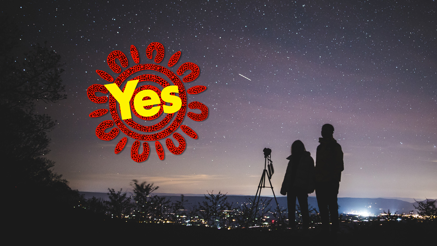 Two people and a camera on a tripod are in silhouette against the night sky. In the near distance, the lights of a small city or town sparkle between black trees in silhouette with mountains further in the distance. The night sky is filled with stars sprinkled on the backdrop of soft hues of dark greys and purples. Overlaid on the photo is the red and yellow icon of the 'Yes' campaign.