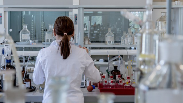 A scientist in a lab coat facing a lab bench with her back to the photographer. A fume cupboard is in the distance.