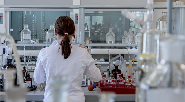 A scientist in a lab coat facing a lab bench with her back to the photographer. A fume cupboard is in the distance.